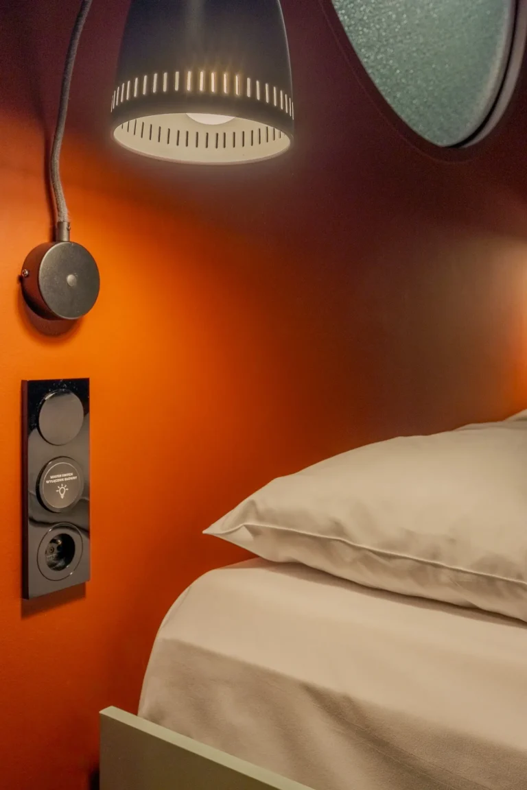 Miękkie światło z lampki do czytania pada na łóżko i czerwoną ścianę.