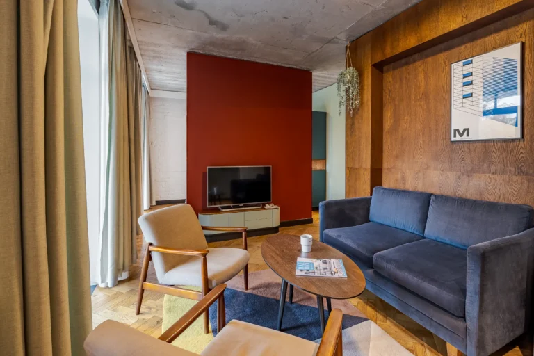 Pokój hotelowy typu studio z drewnianym parkietem, niebieską sofą i czerwoną ścianą.