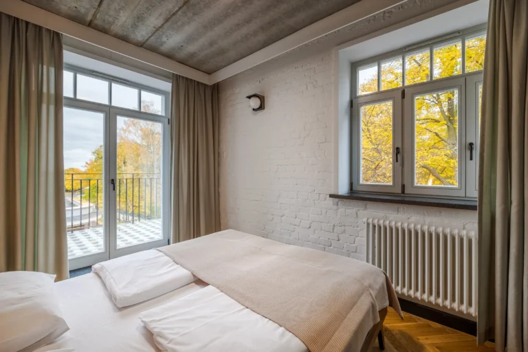 Pokój hotelowy z podwójnym łóżkiem i jesiennymi drzewami za oknem.