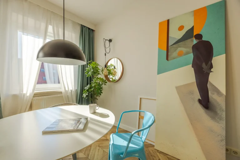 Duży nowoczesny obraz stoi obok okrągłego stołu i niebieskiego krzesła.