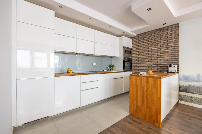 Biała, jasna kuchnia z drewnianymi blatami, wyspą i ceglaną ścianą.